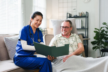 Asian female nurse in blue scrubs showing a medical folder to a smiling senior man during a home healthcare visit in a living room.