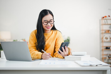Young Asian woman with glasses smiling while using a smartphone and holding a pen at a white desk with a laptop in a bright home office.