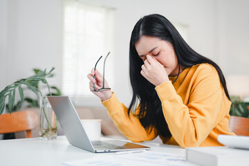 Asian woman rubbing her eyes and holding glasses while working on laptop, feeling tired, stressed, and suffering from digital eye strain or headache in home office.