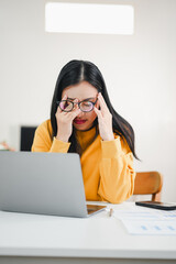 Exhausted young Asian woman with glasses holding her head and feeling stressed while working on her laptop at a desk in a home office.