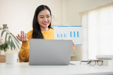 Young Asian woman in yellow sweater presenting a business sales report chart during a video call on a laptop in her home office.