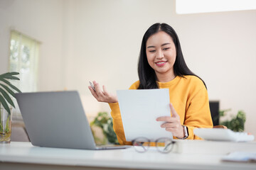 Smiling Asian woman in yellow sweater looking at document while working at home office desk with laptop and calculator.
