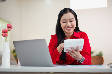 Smiling Asian woman in a red shirt using a calculator while working on a laptop at her home office desk.