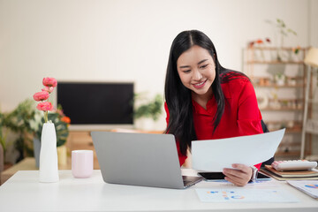 Young Asian woman smiling while reviewing paperwork and working on a laptop at a home office desk.
