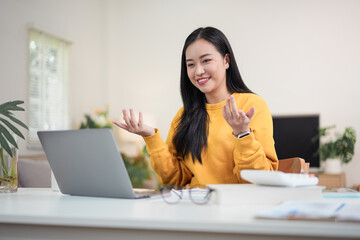 Happy young Asian woman gesturing and smiling while having a video call on a laptop in her bright home office during remote work.