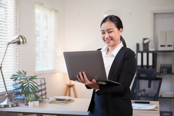 Professional smiling Asian businesswoman in a black suit holding a laptop and working in a bright modern office environment.