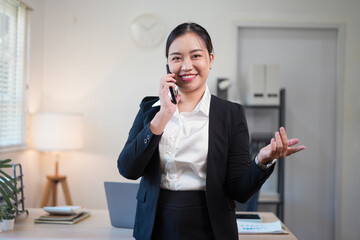 Smiling Asian businesswoman in a professional black suit talking on a smartphone and gesturing while standing in a bright modern office workspace.