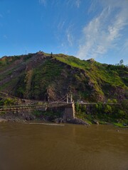Scenic suspension bridge crossing a wide river at the foot of a lush green mountain under a clear blue sky with wispy clouds.