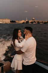 Fototapeta premium Beautiful young couple embraces and kisses on yacht cruising European city canals at sunset warm twilight colors romantic flowers joyful moment love and intimacy cozy evening atmosph