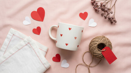 White mug with hearts, red paper hearts, and ball of yarn on pink fabric