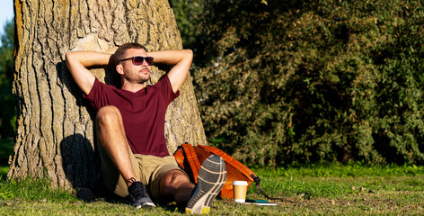 Urban man relaxing in city park, sitting near tree with hands behind head.