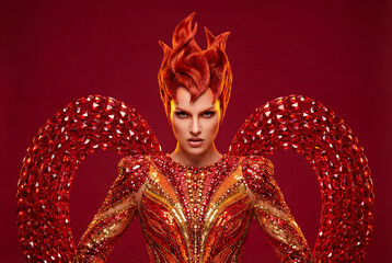 A fierce woman personifies a mythical phoenix or firebird. An elaborate red and gold sequin costume, flame-like hair, and a powerful, confident gaze.