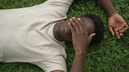 Young African American man lying on grass, covering face with hand, embracing nature, experiencing digital detox and relaxation in a serene outdoor environment