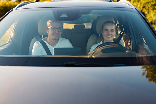 Two people are inside a car, smiling and driving along a sunny road lined with trees. Bright sunlight shines through the windshield as they enjoy the ride