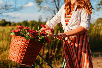 A person stands next to a bicycle with a basket filled with flowers in a field during sunset. The...