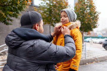 African American man taking care and dressing his son in warm jacket and hat outdoors, father buttoning his child's shirt and checking warm clothes in autumn in the city