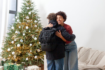 African-American woman hugs her son and gets him ready for school at home near the Christmas tree, teenager gets ready for school in winter with backpack and says goodbye to his mother