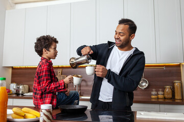 African American man and his son drink coffee in the kitchen in the morning and talk, teenager and his father drink tea at home and communication