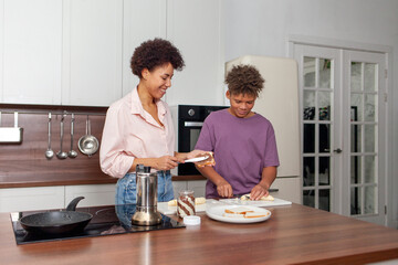 African-American woman and her son prepare chocolate butter and banana sandwiches in the kitchen, while teenager helps his mother prepare breakfast at home.