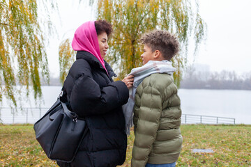 African American woman and her son are walking in the park in autumn wearing warm clothes, the mother is taking care of the child and putting scarf on the boy