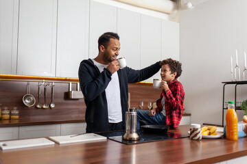 African American man and his son drink coffee in the kitchen in the morning and talk, teenager and his father drink tea at home and communication