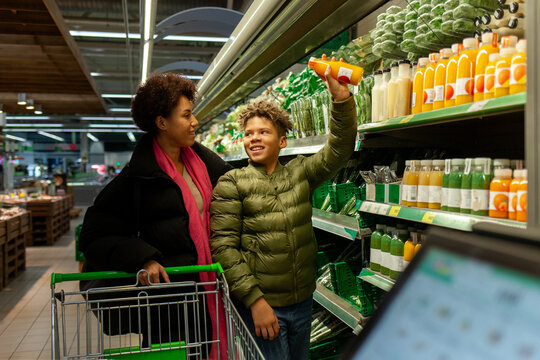 African American woman and her son shop for groceries at supermarket, teenager chooses orange juice and shows it to his mother in the store