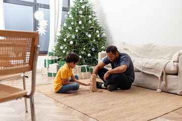 African American man and his son play board game at home against Christmas tree, father and child build tower of wooden blocks in educational game