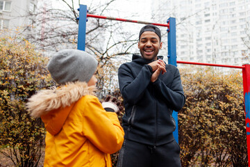 African American man and his son exercise and do exercises outdoors on playground, teenager stretches with his father on exercise equipment