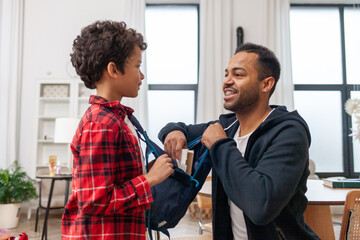 African American man gets his son ready for school, puts jacket and backpack on him at home, and the father takes care of the child and puts book in the backpack