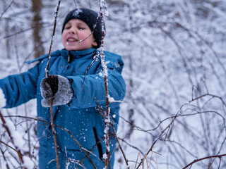 Smiling Boy Exploring Snow-Covered Branches in a Winter Forest - Winter Nature Walk: A Child in a Blue Jacket and Black Beanie Playing in the Snow