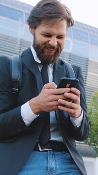 Portrait of man office worker with well-groomed beard in black jacket and jeans which laughing from revision amusing pictures on mobile while stopped on bike on the big urban building background