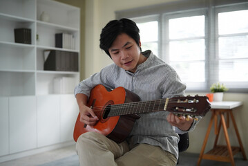 A young Asian man sits comfortably at home, practicing acoustic guitar in a calm, naturally lit living space