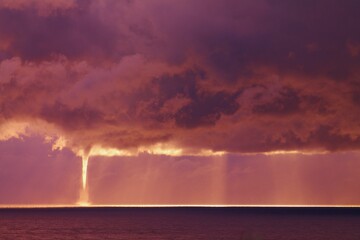  A waterspout is visible beneath thick, dark clouds over the ocean, stretching down from the sky towards the water. Sun rays peek through the cloud cover, illuminating parts of the sea.