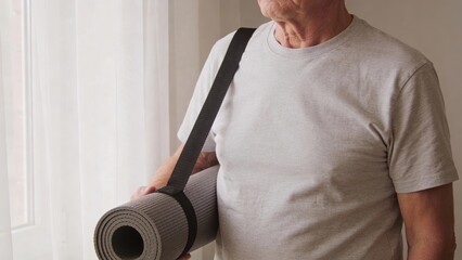 Older man holding a rolled yoga mat by a sunlit window Retirement Living concept