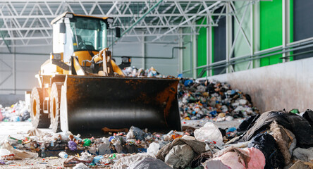 Industrial bulldozer in waste management facility sorting recyclable materials and trash to conveyor of garbage recycling plant