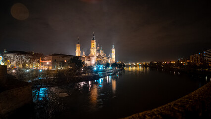 Fototapeta premium Dynamic wide-angle night shot of the Basilica del Pilar in Zaragoza, with its illuminated towers and domes beautifully reflected in the flowing waters of the Ebro River during the Christmas season.