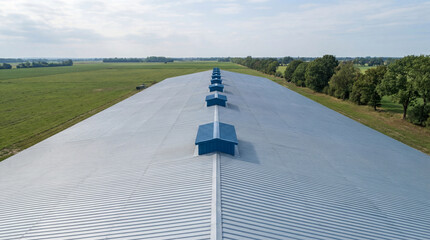 High-angle aerial view of a long industrial metal warehouse roof with blue ventilation skylights in a rural green field.