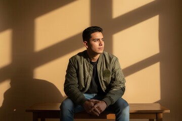 A pensive young Latino man in a bomber jacket sitting in direct sunlight. Thoughtful Hispanic male model posing indoors with dramatic shadows on a beige wall. Modern contemplative portrait