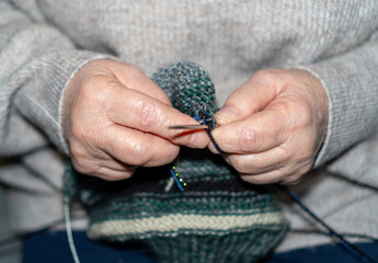 A detailed macro shot of a senior woman's hands knitting a garment using dark green melange wool yarn and metallic needles. The focus is on the intricate movement of the fingers and the texture of the