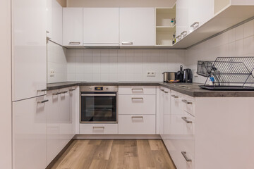 Horizontal wide angle eye level shot of a clean L-shaped kitchen featuring white glossy cupboards, built-in oven, and a wooden floor in a studio