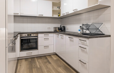 Horizontal wide angle eye level shot of a clean L-shaped kitchen featuring white glossy cupboards, built-in oven, and a wooden floor in a studio © Igor