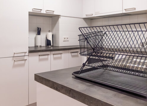 Horizontal wide angle eye level shot focusing on a sleek metal dish rack in a minimalist white kitchen with blurred cabinets and utensils behind