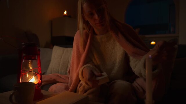 Young woman snuggled in blanket at home during power outage, lighting candles with match and enjoy book by soft glow of kerosene lamp in warm and dimly lit room