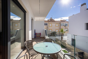 Sunny residential balcony with glass table, chairs, and clear sky view over rooftops