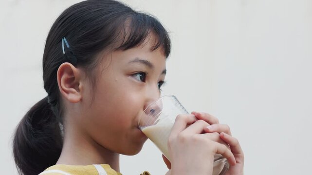 Close up of adorable Asian girl drinking fresh milk from glass in natural light. Cute child holding milk glass enjoying healthy breakfast nutrition and calcium for growth and development.