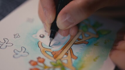 Close up of fingers outlining a miniature ghost cat watercolor painting with an ink pen. Whimsical art process on paper, shallow depth of field, focused on pen detail, sketching and creative hobby.