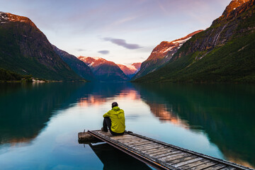 Man sits on wooden jetty looking at mountain reflection in lake in Norway during sunset