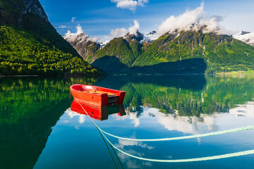Red boat floats on still lake in Norway with mountains and clouds in the background during sunny day
