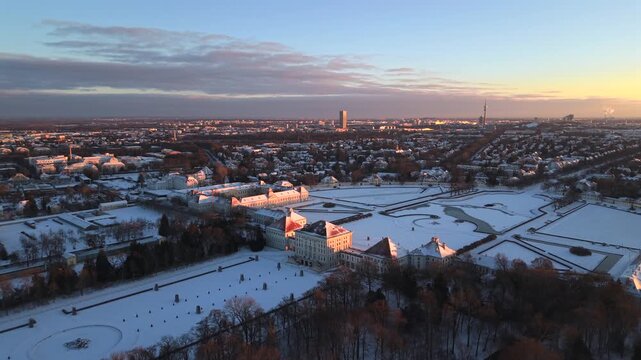 Cinematic aerial view of Nymphenburg Palace in Munich, Germany. Winter dawn with fresh snow highlights the historic baroque palace, frozen canals and royal gardens in calm morning light.