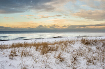 Snow-covered dunes with dry grass overlooking calm Baltic Sea at sunset.